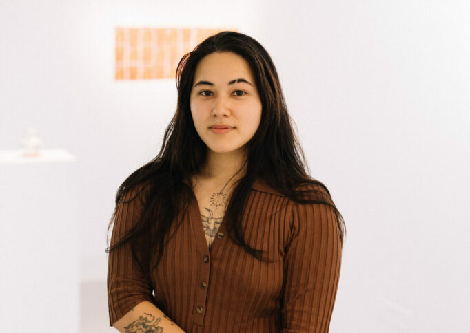 Portrait of a person with long straight brown hair, a rust brown cardigan, chest and arm tattoos, and a shaved eyebrow slit.