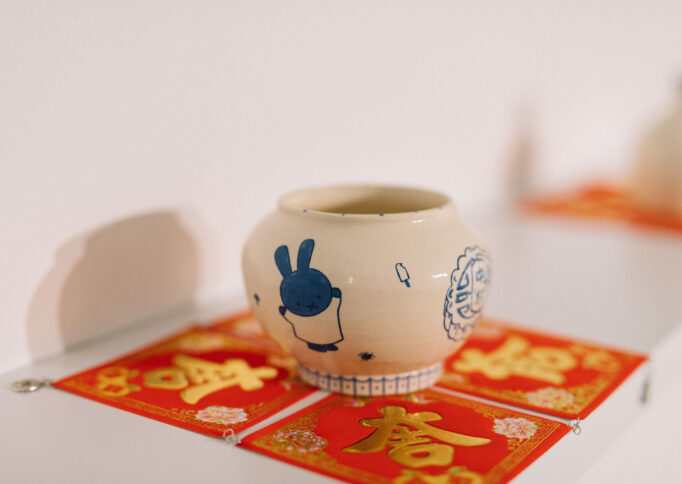 Detail of a cream ceramic vessel with blue image of a rabbit, presented on red square fabric, on a floating white shelf.