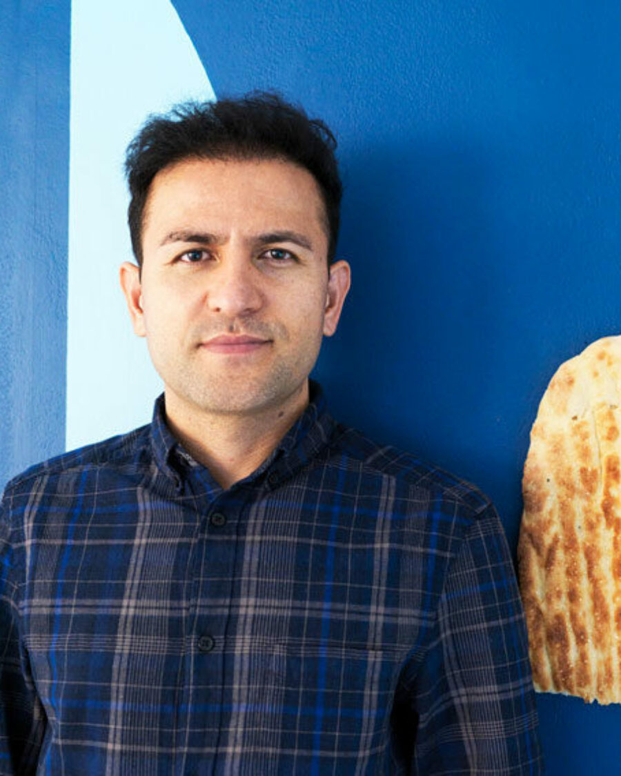 Portrait of Elyas Alavi in a navy check shirt against a blue wall. There is half a length of Afghan bread on the wall.