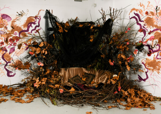 A shrine like sculptural installation in the corner of the gallery of dried flowers, foliage, black sheer fabric and silken gold fabric against an abstract wall painting.