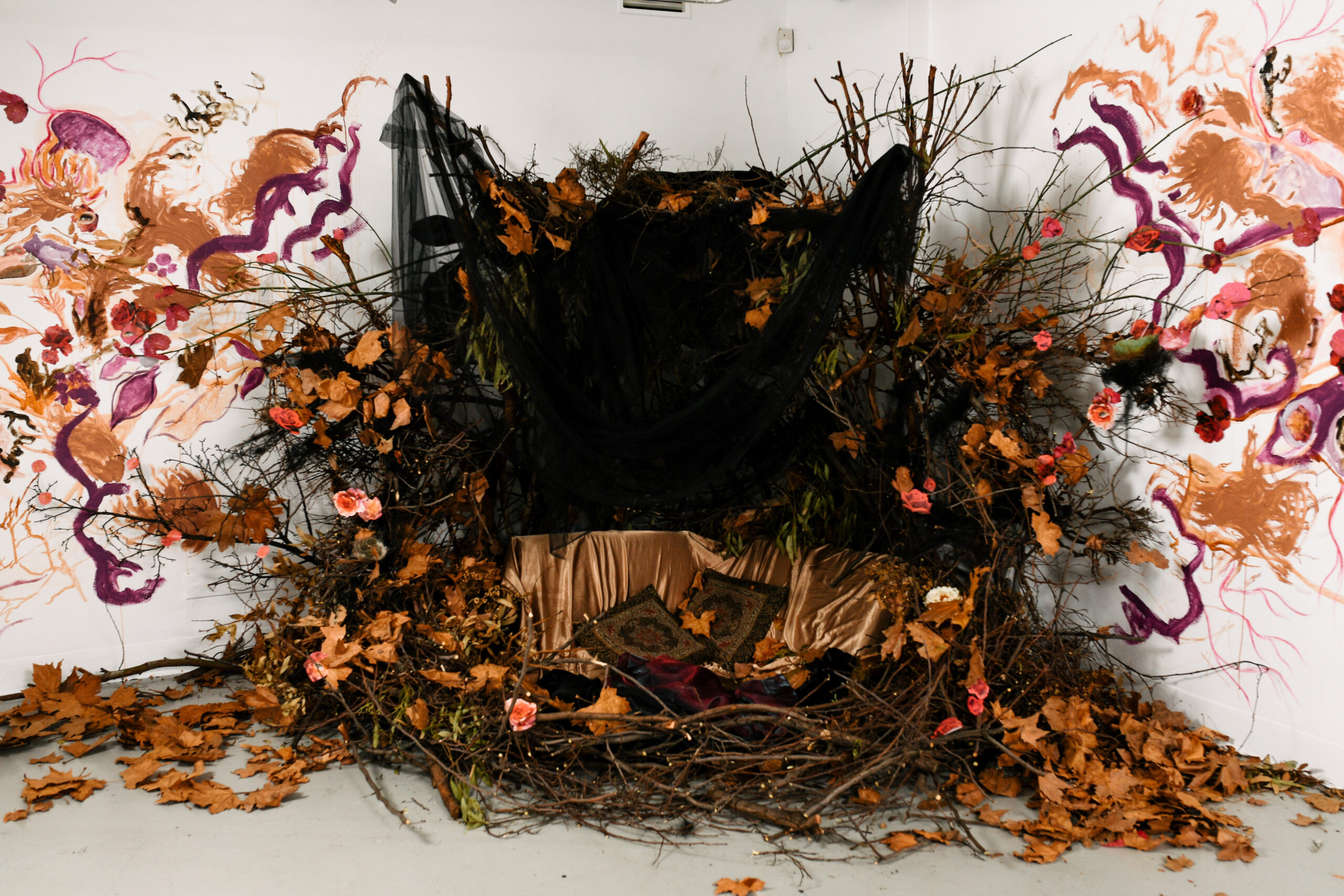 A shrine like sculptural installation in the corner of the gallery of dried flowers, foliage, black sheer fabric and silken gold fabric against an abstract wall painting.
