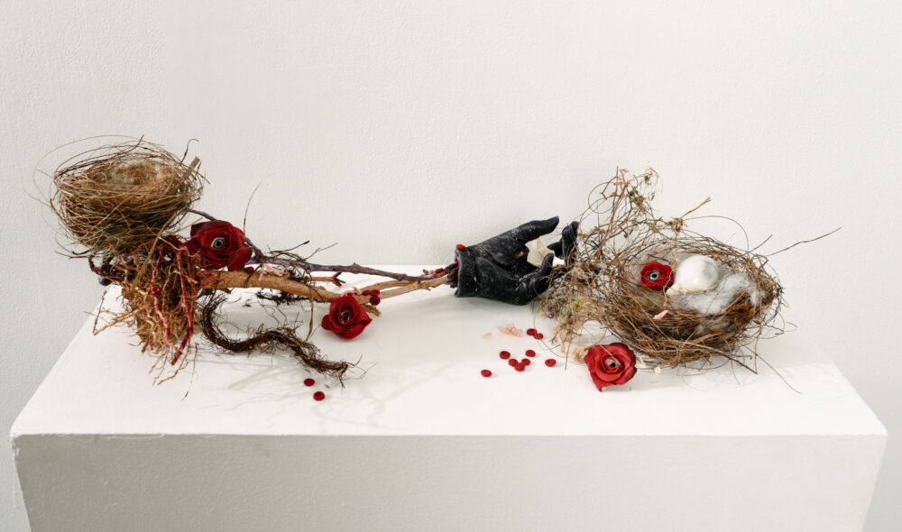 Detail of sculptural work on a white plinth. The sculpture is composed of 2 nests, a black hand, flowers and twigs.