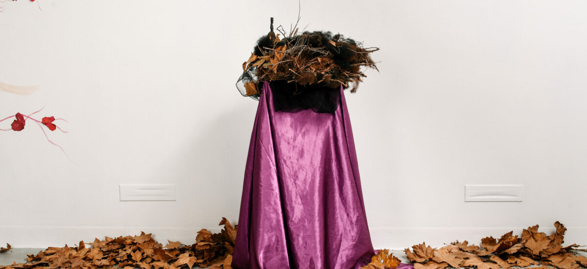 Sculpture of a nest made from dried foliage, set on a pink silk fabric covered plinth. There is a white and black egg in the nest and dried autumn leaves on the floor.