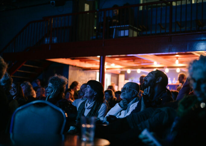 In a dark blue light, a seated audience with different expressions all look in the direction of the stage.