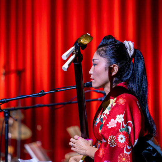 Profile of Noriko Tadano playing a shamisen on stage in front of a red curtain, she wears a floral red kimono, red lipstick and has a long black ponytail secured with a scrunchie.