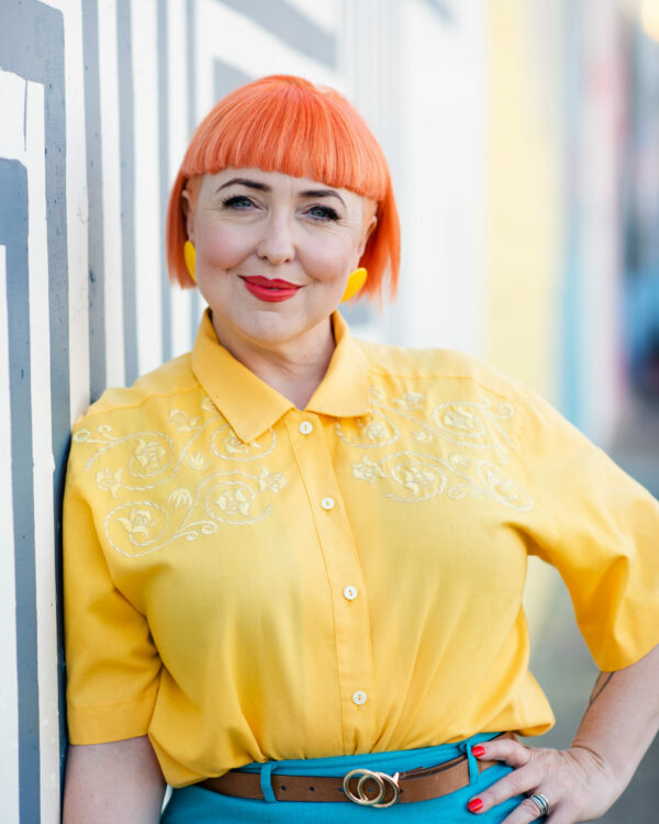 Headshot of Peta-Anne, leaning against a wall wearing a bright yellow shirt, button yellow earrings and a warm smile. She has orange bob hair with a blunt fringe.