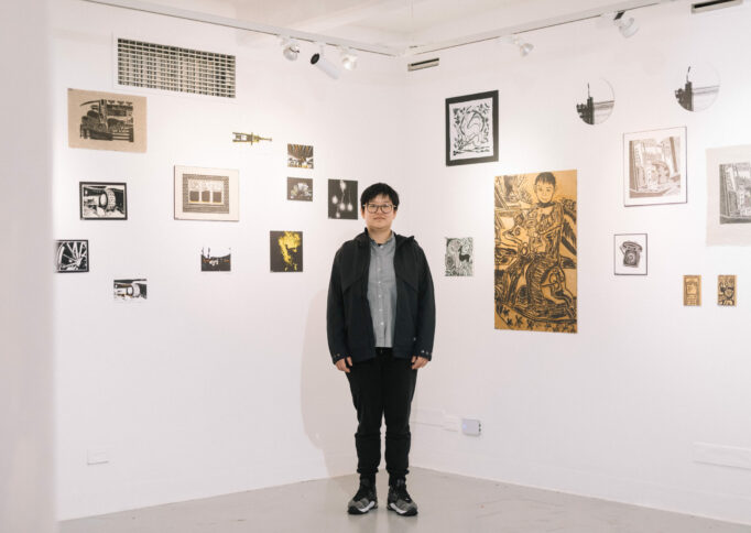 A person with short black hair and glasses stands in front of a salon hang of small artworks.