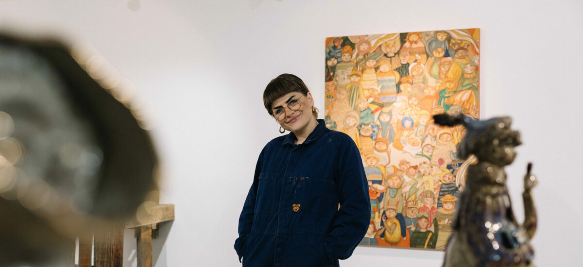 Portrait of arist Fruzsi Kenez standing amidst an installation. Fruzsi has a short fringe, dark eyebrows, glasses and is wearing a navy boiler suit.