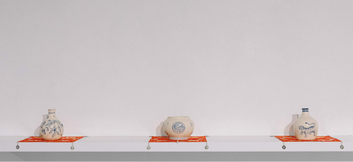 Three cream ceramic vessels of different shapes with blue images on them, presented at equal distances on red square fabric, on a floating white shelf.