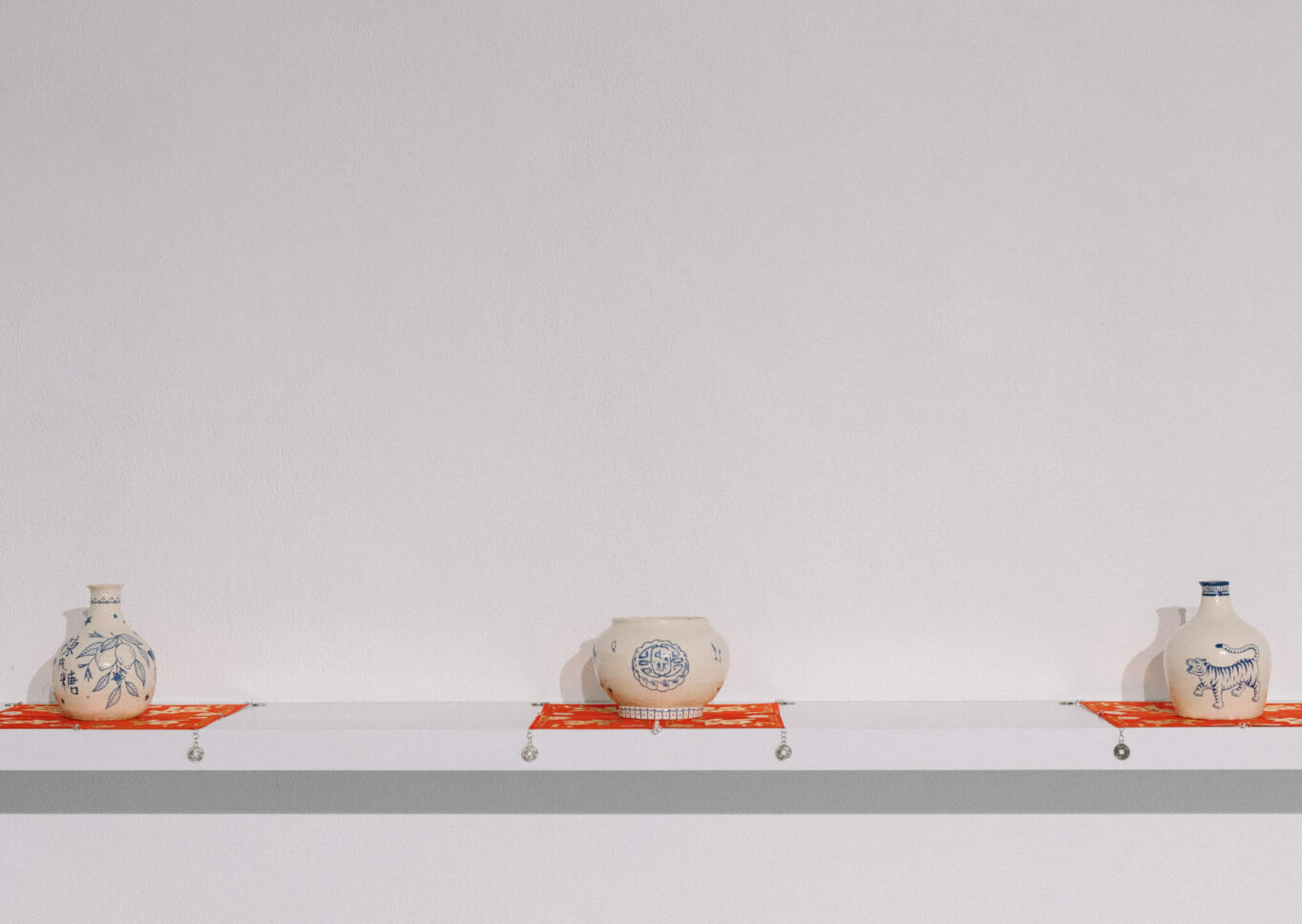 Three cream ceramic vessels of different shapes with blue images on them, presented at equal distances on red square fabric, on a floating white shelf.