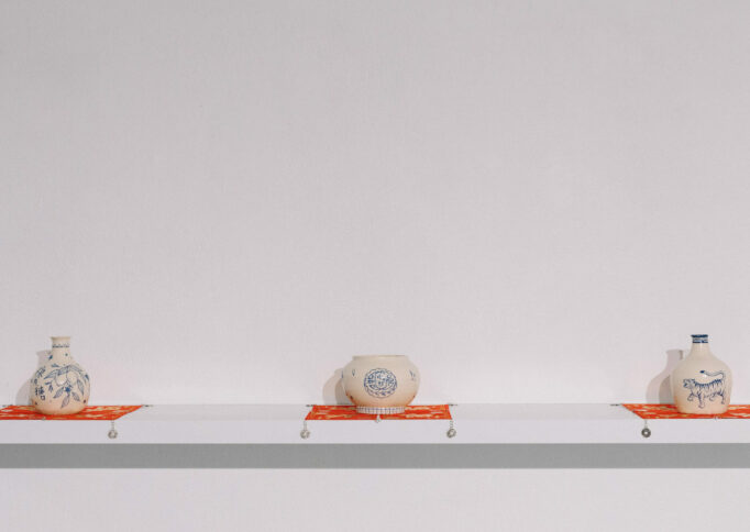 Three cream ceramic vessels of different shapes with blue images on them, presented at equal distances on red square fabric, on a floating white shelf.
