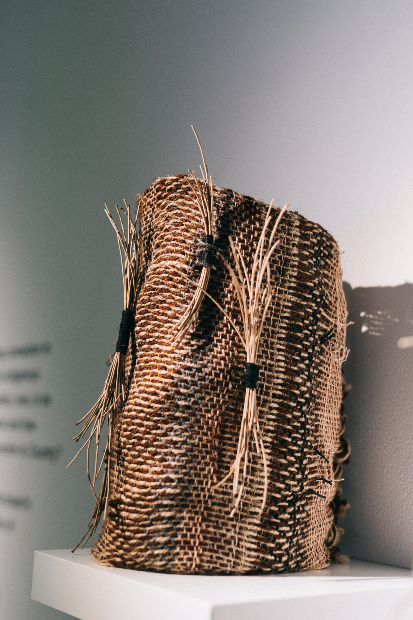 A cylindrical vessel made from jute, recycled wool, reeds, sheoak nettles, presented on a floating white shelf.