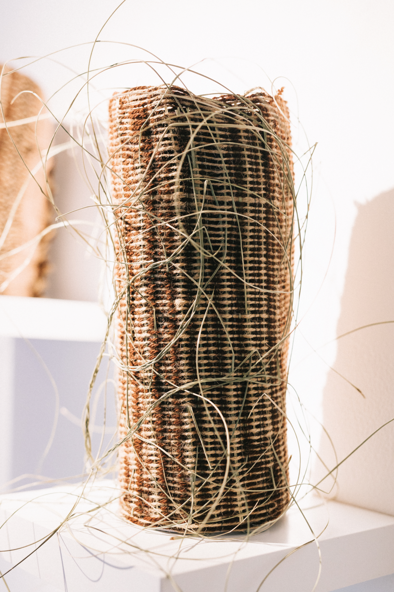 A tall vertically presented vessel made from jute, recycled wool, reeds, sheoak nettles, presented on a floating white shelf.