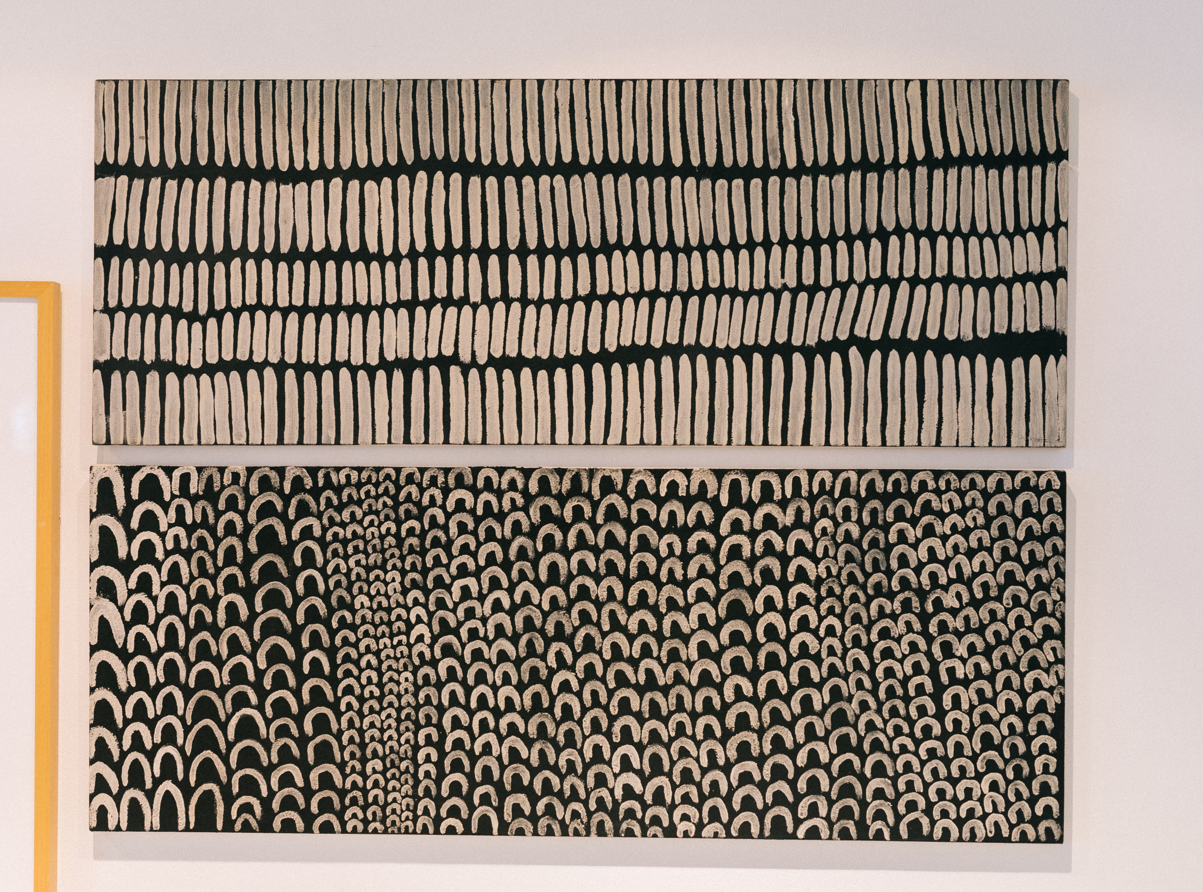 Two black and white paintings with a horizontal orientation. One work depicts many vertical lines in five rows, the other painting features repeated horseshoe shapes.