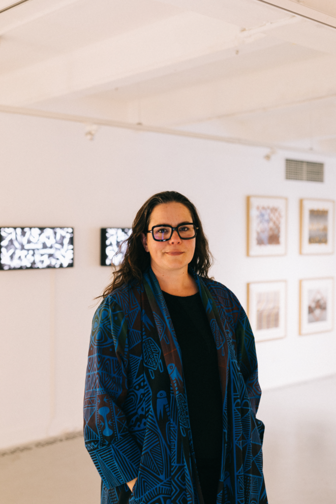 Portrait of a person with long dark hair and dark framed glasses. They stand in front of a wall of artworks with their hands in their pocket