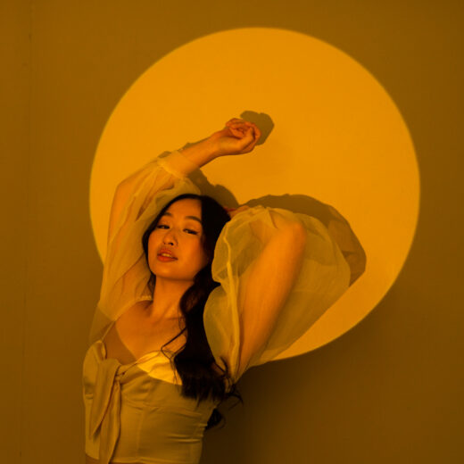 Elizabeth Ruyi wears a white dress with ballooning sheer sleeves, leaning back, arms above her head, lit in a warm yellow circular spotlight.