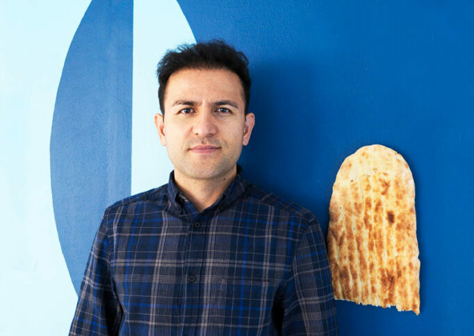 Portrait of Elyas Alavi in a navy check shirt against a blue wall. There is half a length of Afghan bread on the wall.