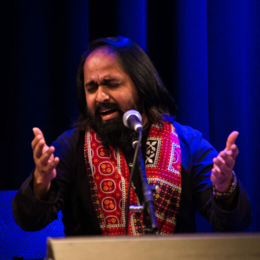 A man with medium long brown hair, moustache and beard holds his open hands palms up, while singing with eyes closed into a microphone.