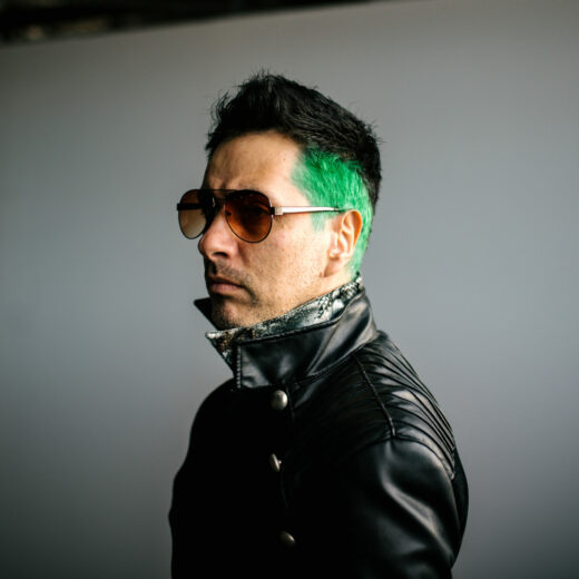 Side profile of a person with a short brown and green Mohawk hairstyle, aviator glasses on wearing a black leather jacket with popped collar.