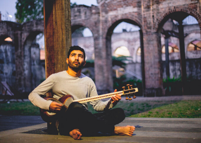 A man playing a string instrument, sits on the grass, legs crossed and back against a pole, looking up into a warm yellow light.