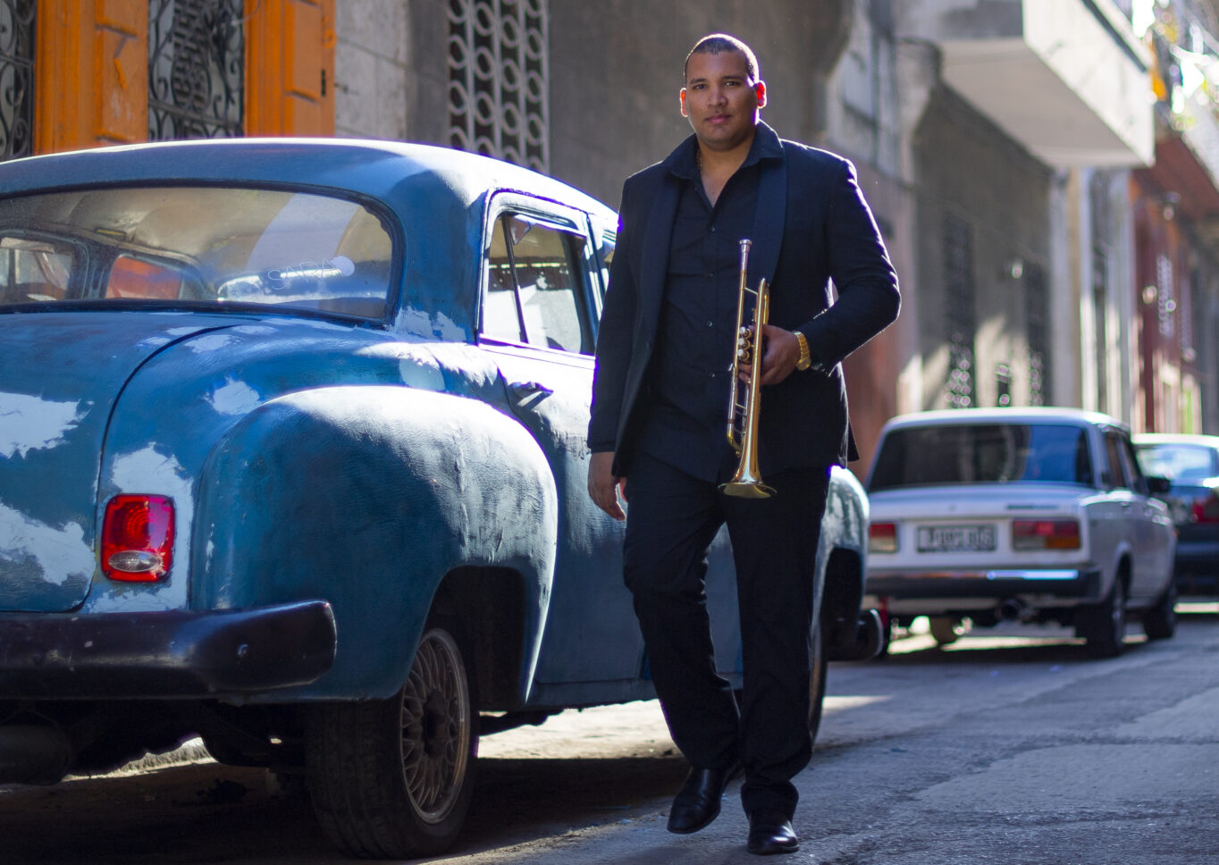 Trumpeter Lazaro Numa walking through a city street in a black suit holding his trumpet.