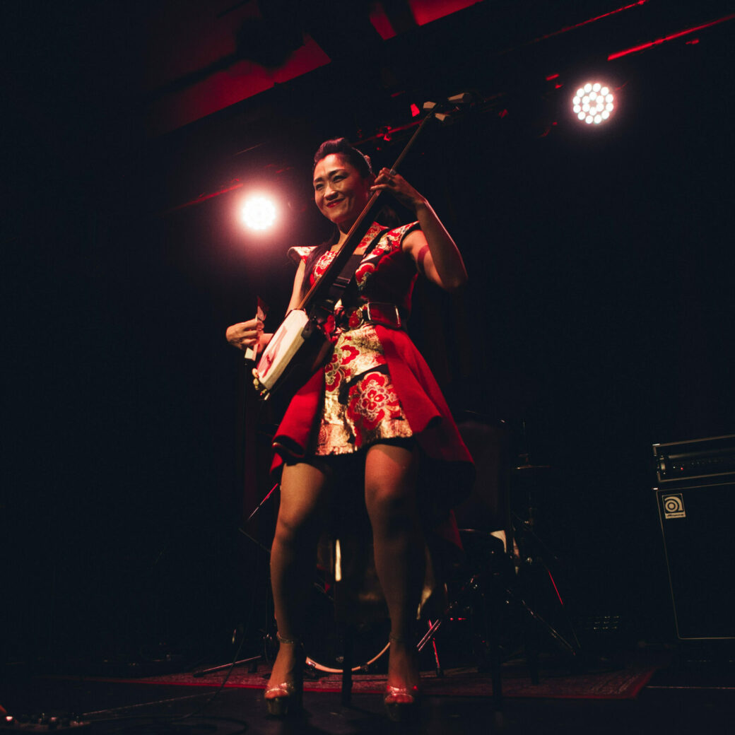 On stage, a woman in a short floral red dress and red heels plays a shamisan, she is smiling.