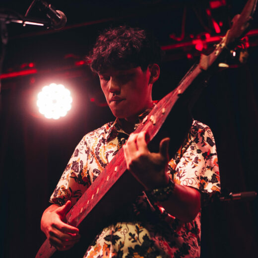 Lit in red, a man with short dark curly hair and patterned shirt plays ornate string instrument, a sampe’ on stage.