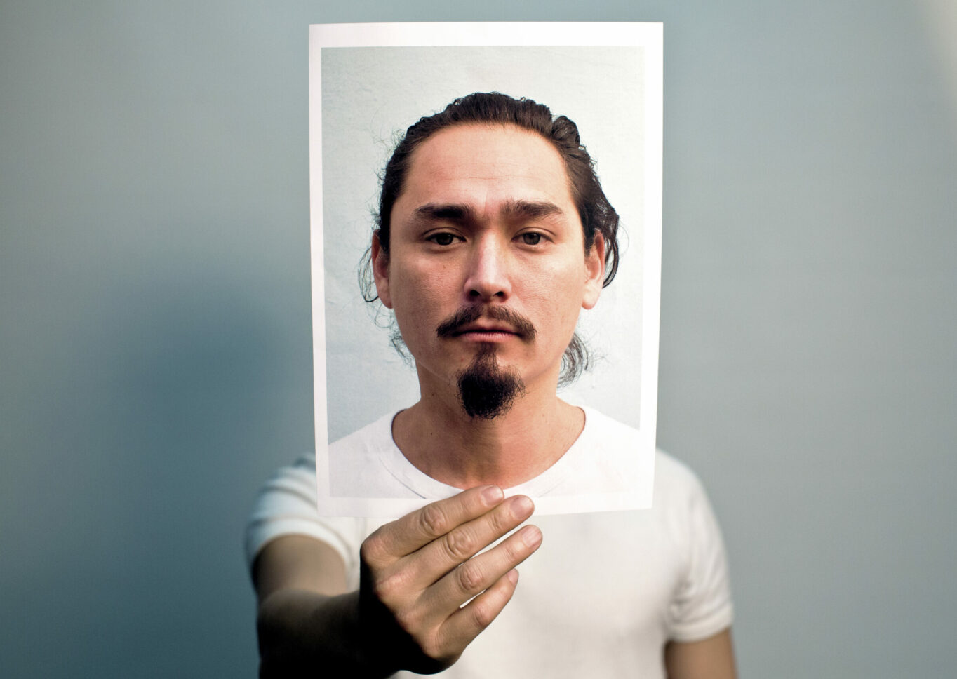 A man in a white t-shirt holds up a photograph of his face out in front where his head would be.