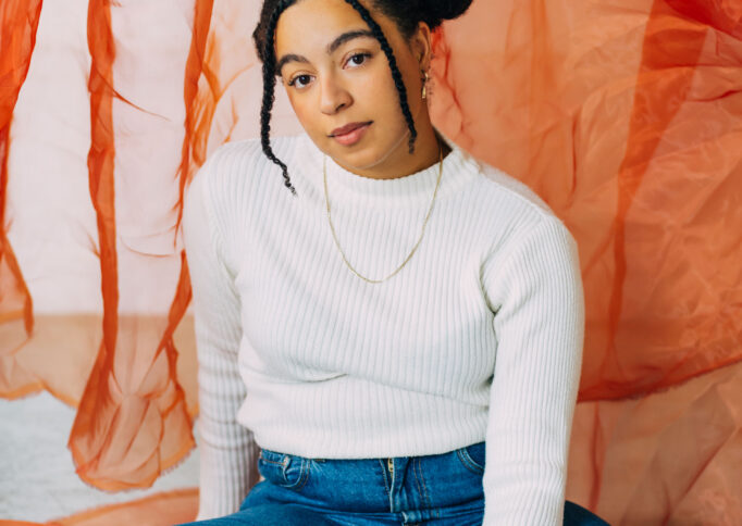 A photograph of Kayla. She wears a white top blue jeans and is sitting in front of an orange screen.