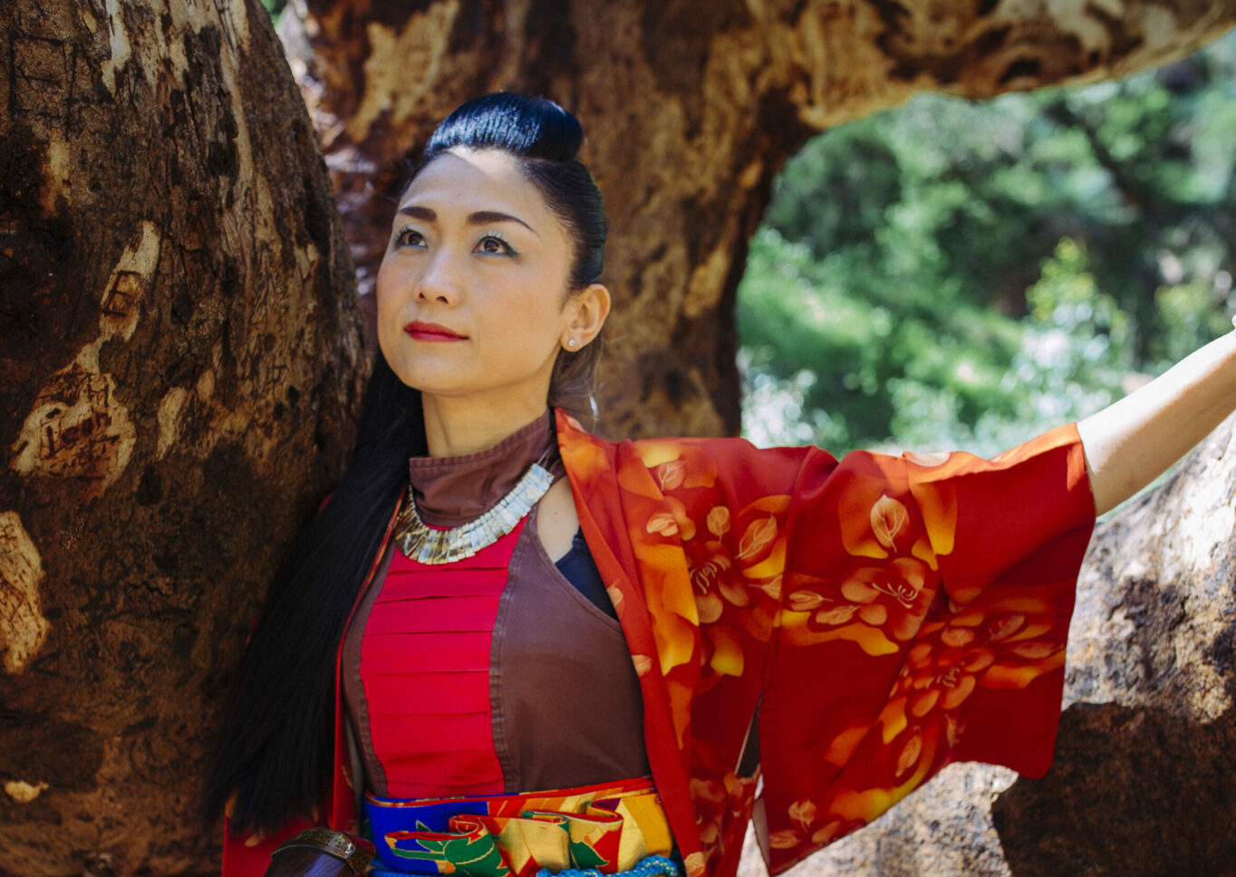 Portrait of Noriko Tadano in nature near a big tree trunk. She wears a traditional Japanese Kimono and holds her instrument the shamisen.