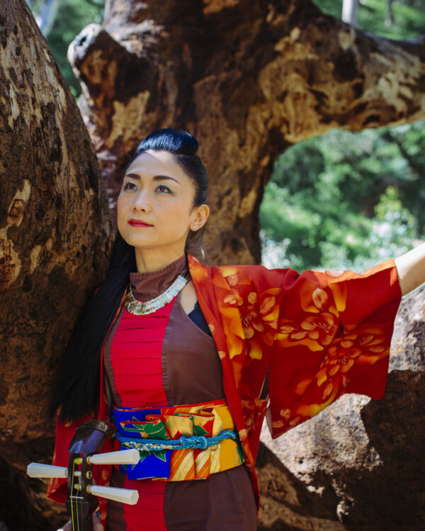 Portrait of Noriko Tadano in nature near a big tree trunk. She wears a traditional Japanese Kimono and holds her instrument the shamisen.