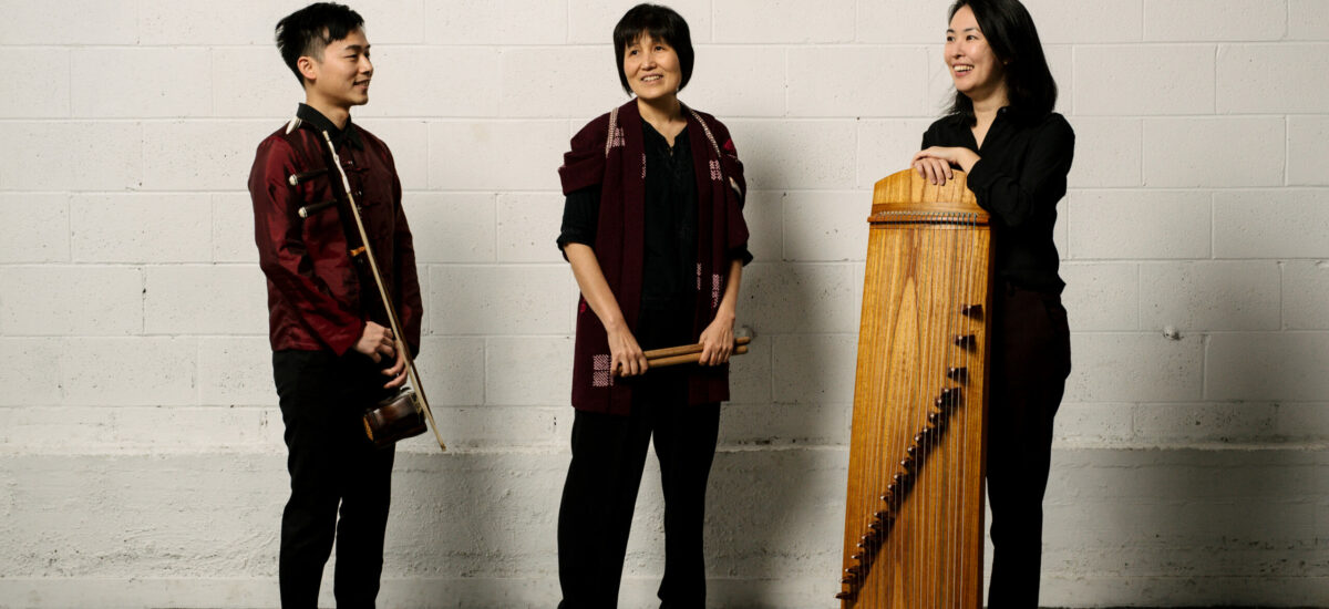 San Ureshi musicians wearing black and maroon outfits, stand posed with their instruments in front of a white brick wall.