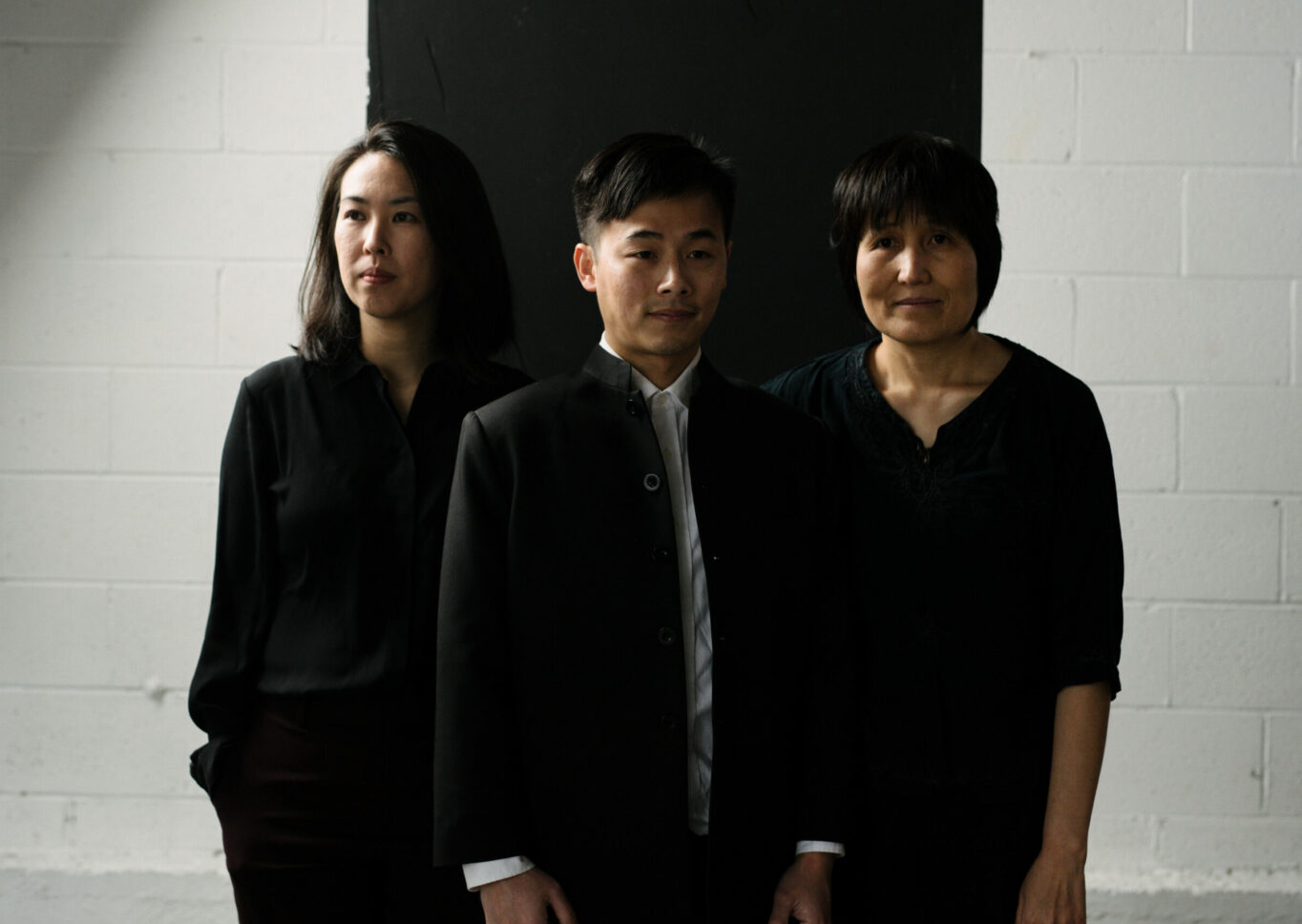 San Ureshi band members- three people all dressed in black, stand posed in different angles in front of a white brick wall.