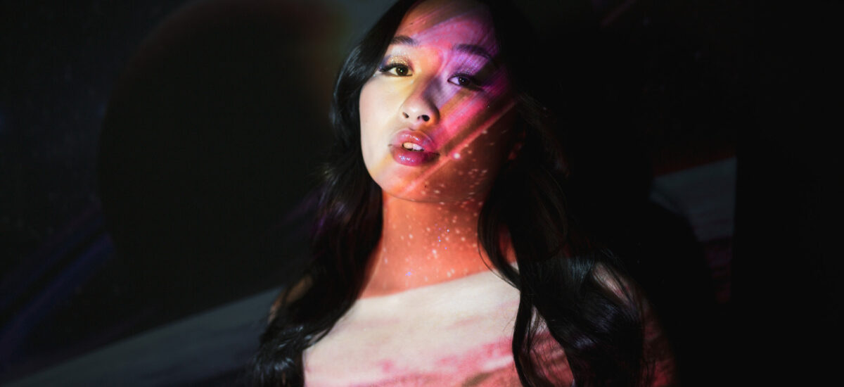 A portrait of a woman with long dark hair and open lips is covered by galaxy projections across her face and chest.
