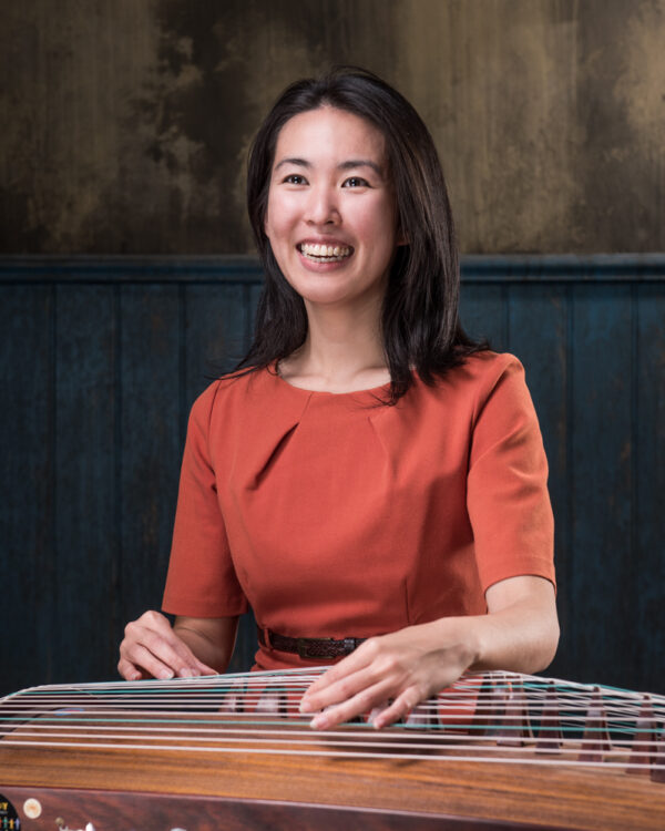 Portrait of Zhao Liang smiling. She wears a red top and plays an instrument.