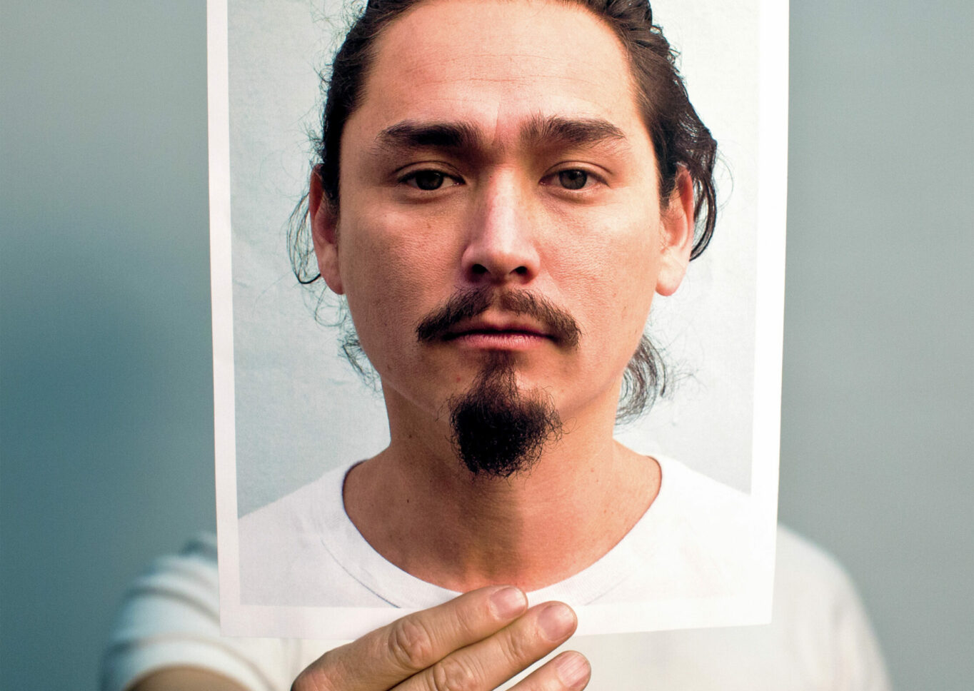 A man in a white t-shirt holds up a photograph of his face out in front where his head would be.