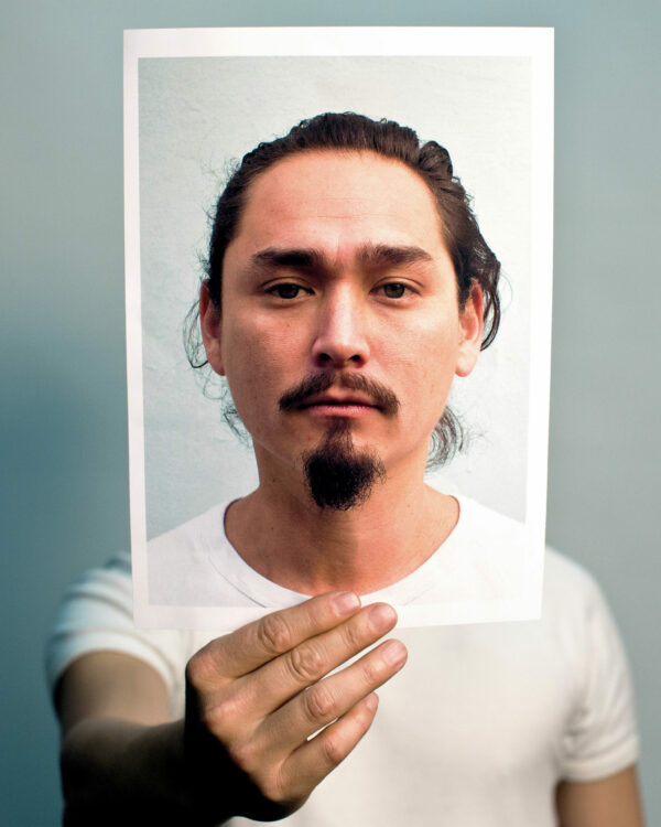 A man in a white t-shirt holds up a photograph of his face out in front where his head would be.