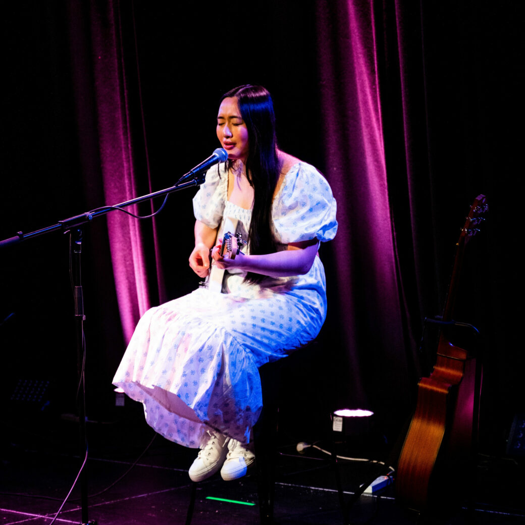On stage a woman in a long white dress and white sneakers sings into a microphone while playing a white electric guitar.