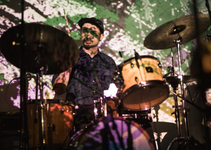 A man in a cap plays the drums, which along with his face and body, are covered with a green and white digital projection.