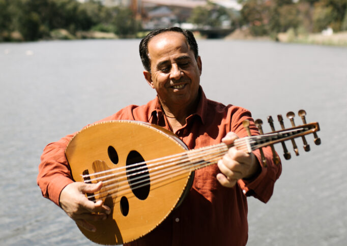 Photograph of musician Zuhir Naji in a rust coloured shirt in-front of a body of water. He is smiling and playing the oud, a short-neck lute-type, pear-shaped, fretless stringed instrument.