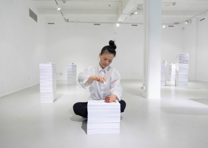 Artist Jingwei Bu sitting in the gallery behind a stack of white paper pouring tea