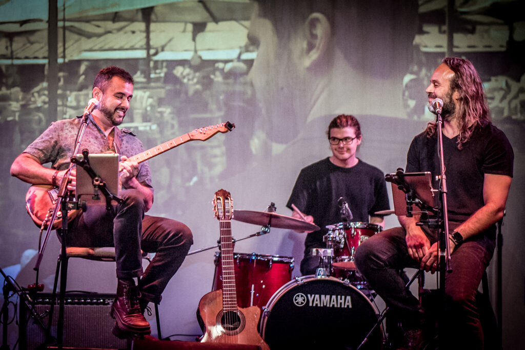 A photograph of three musicians sitting on a stage in front of a projected image. One person is playing guitar and another is sitting behind a drumkit.