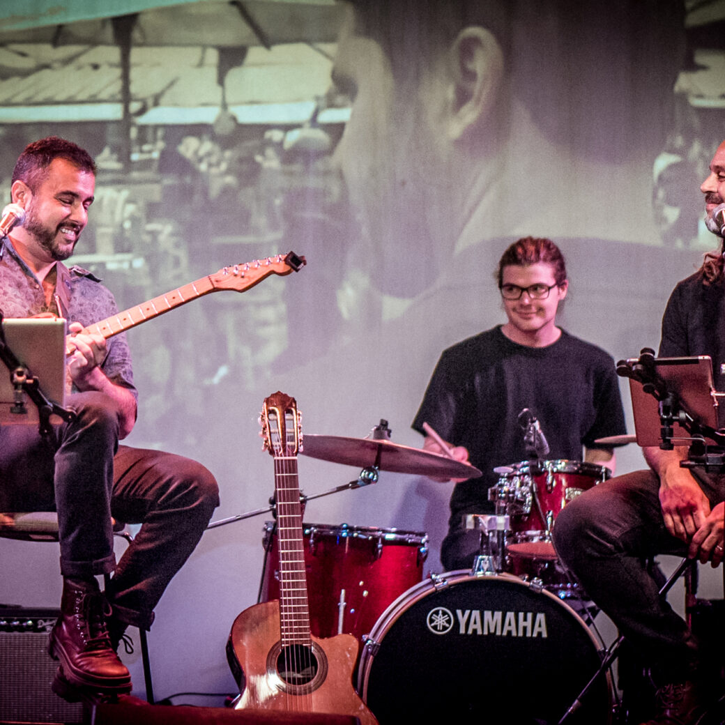 A photograph of three musicians sitting on a stage in front of a projected image. One person is playing guitar and another is sitting behind a drumkit.