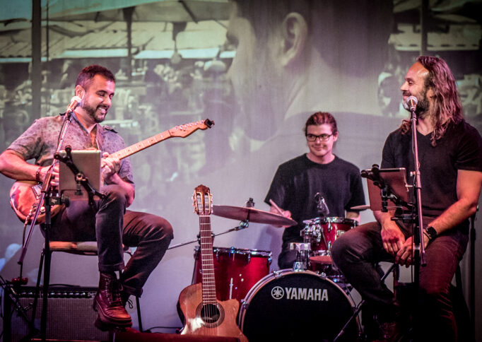 A photograph of three musicians sitting on a stage in front of a projected image. One person is playing guitar and another is sitting behind a drumkit.