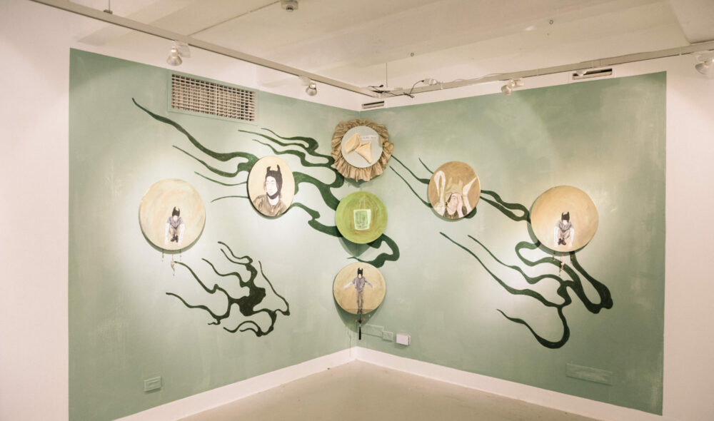 In a corner of the Nexus Gallery, seven circular paintings sit against green background walls with curls of darker green. The paintings feature portraits of a young man, fortune cookies, and Chinese and English text.