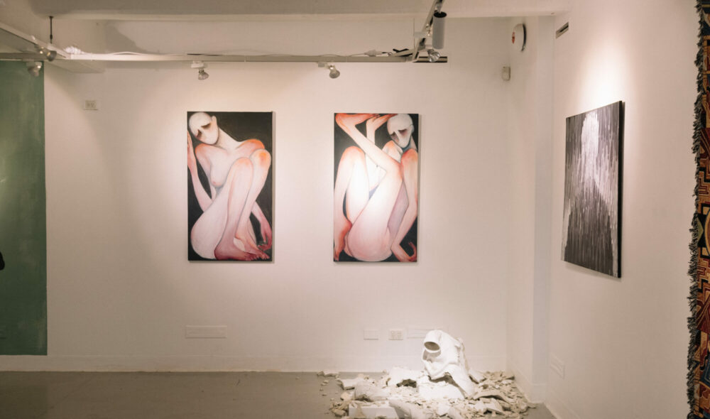 In a corner of the Nexus Gallery, two paintings of contorted figures hang side by side. Below them is a smashed white ceramic work on the floor.