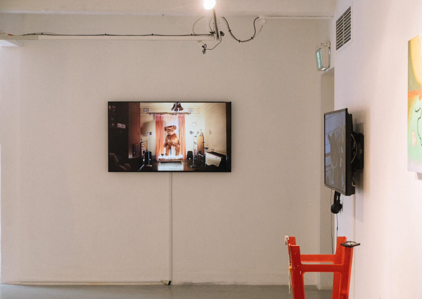 Installation documentation showing a teddy bear on a wall mounted screen and some paintings and sculpture.