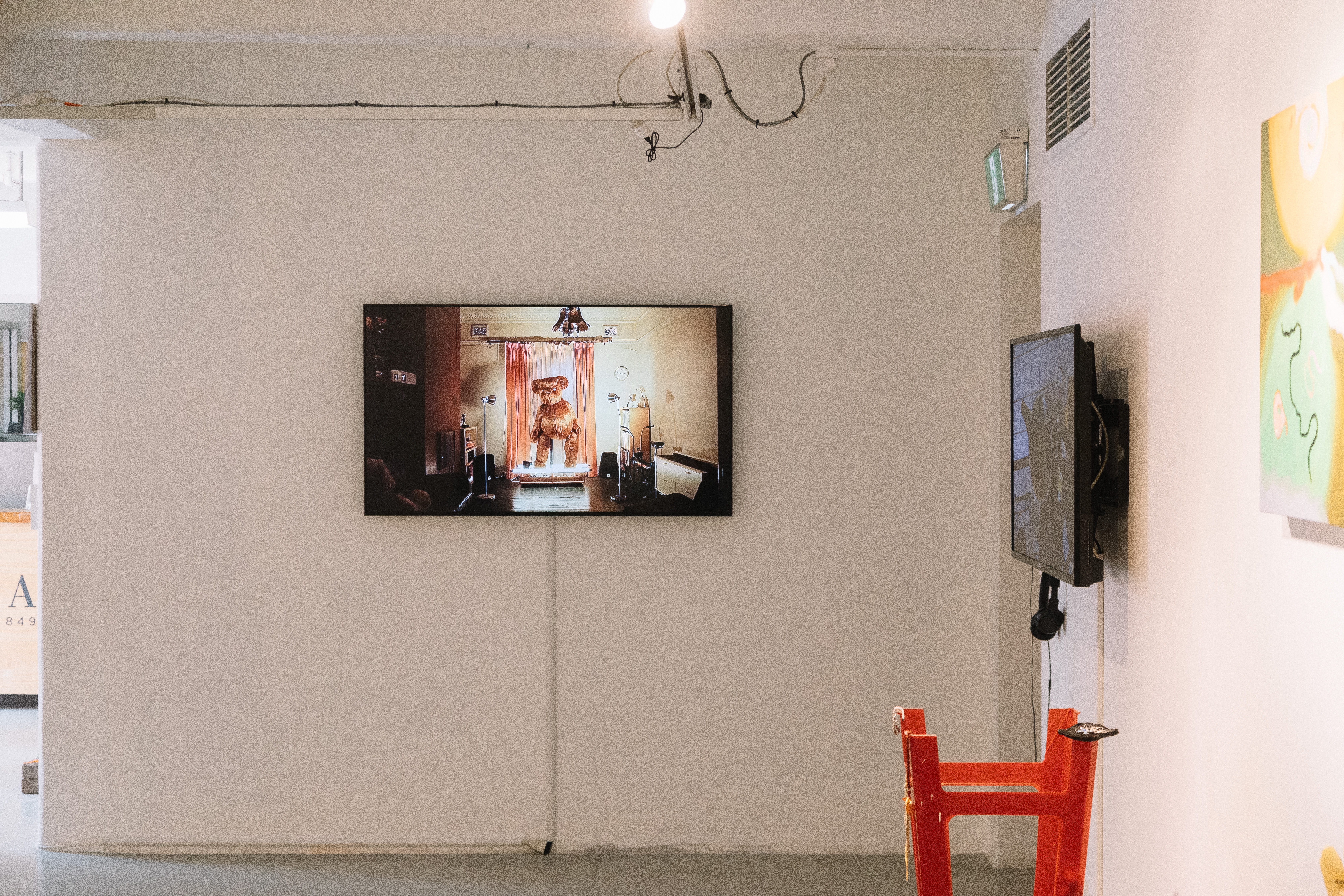 Installation documentation showing a teddy bear on a wall mounted screen and some paintings and sculpture.