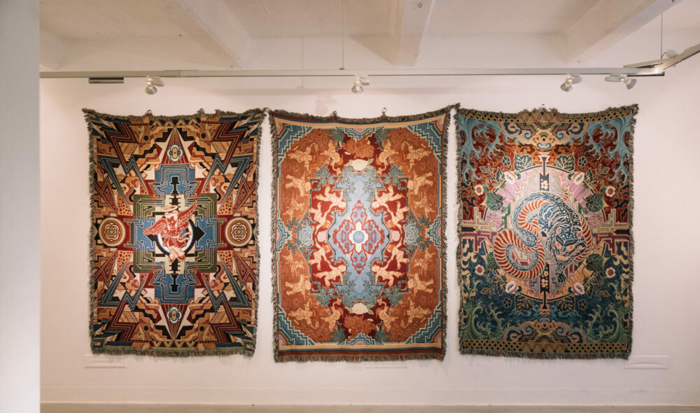 ID: A photograph of three large woven tapestries by [artist]. Each tapestry is around 2m tall and 1.5m wide, and filled with detailed geometric, illustrative designs in brown, red, orange, blue and white. The images include an angelic figure making metal handsigns, crowds of doll-like humans dancing with leaves, and a large blue tiger with a long snake tail. Alt: Three large woven tapestries filled with detailed geometric, illustrative designs in brown, red, orange, blue and white.