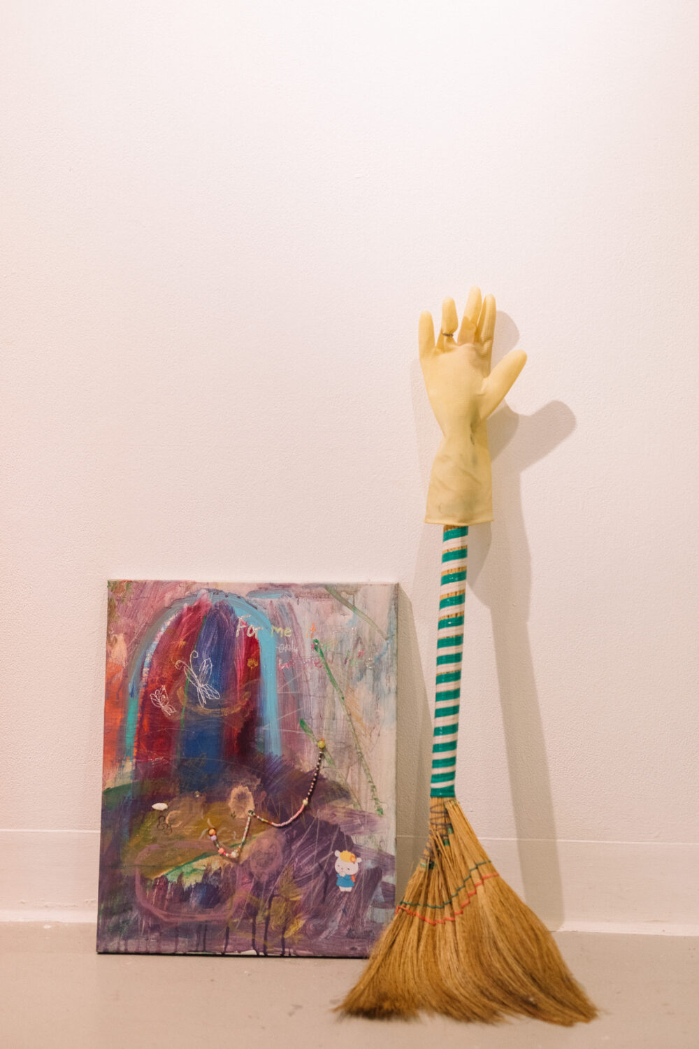 Sitting on the floor of the Nexus Gallery is a 60x40cm painting with abstract purple, red and blue forms, snippets of words, and strings of coloured beads. To the right of this painting is a slightly taller broom, its end capped with a yellow glove.